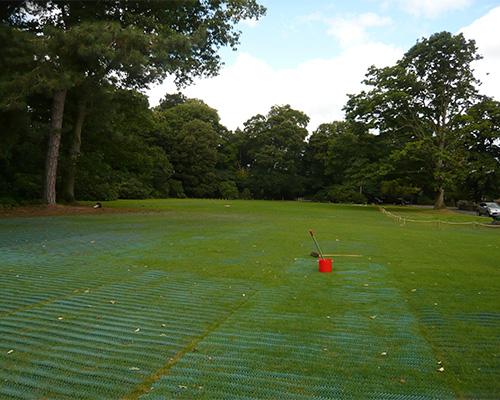 Wide landscape view of Tenax GP Flex ground protection mesh installed across a large event or park field with a building visible in background
