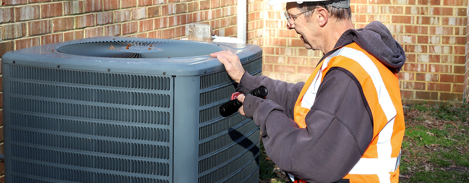 HVAC technician in high-visibility vest servicing a central air conditioning unit at a residential property, showing professional ground protection service context