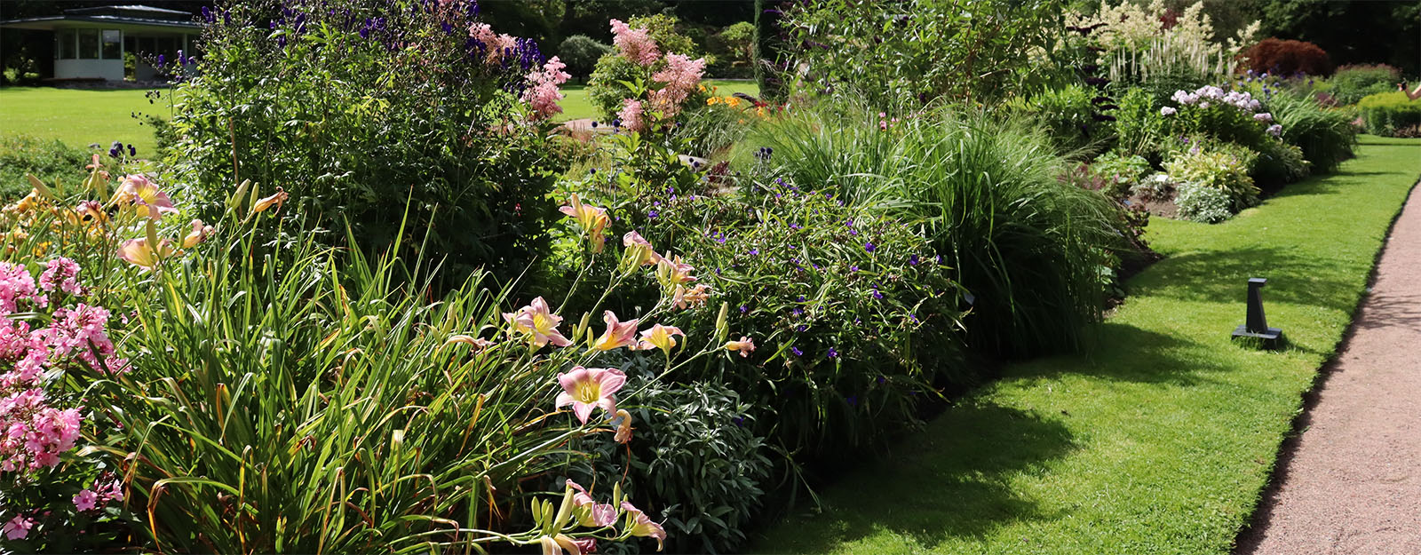 Lush ornamental garden with colourful flower borders flanking a manicured grass path on a sunny day, showing high-quality landscape and lawn maintenance