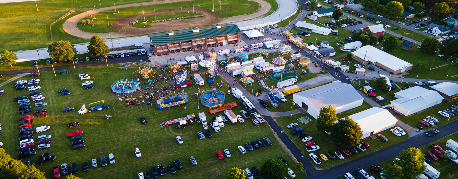 Aerial drone view of a large outdoor festival or county fair showing event tents, crowds, fairground rides, and overflow parking fields requiring ground protection