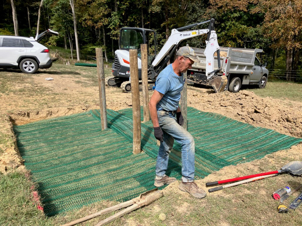 Worker installing Tenax ground protection mesh at a construction site with fence posts being set in reinforced ground